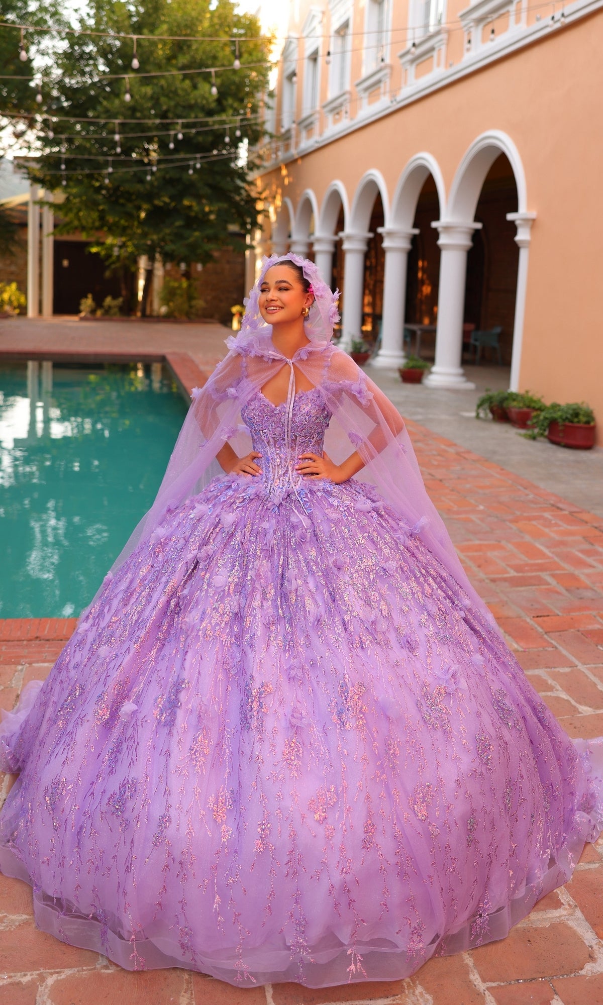 A young woman smiles beside a pool in a courtyard, wearing the Quinceanera Dress 54320 by Amarra?an off-the-shoulder ball gown with sparkly 3D floral details.